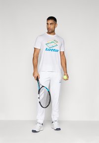 Weißes Lotto-T-Shirt mit buntem geometrischem Logo, kombiniert mit weißen Lotto-Sweatpants. Das Model hält einen Tennisschläger und einen Tennisball.