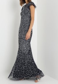 Námořnická modrá večerní šaty s krátkými volánovými rukávy, ozdobené stříbrnými flitry v vlnitém vzoru a průhledným lemem.