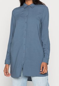 Chemise à manches longues et boutons en tissu bleu atténué. Équipe d'un col classique, coupe droite et légers fentes sur les côtés à l'ourlet.