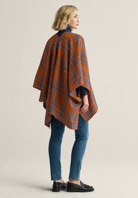 Orange ternet poncho lavet af et blødt stof, med et løst design, der har ærmer i albuelængde og en høj-lav kant. Båret med blå jeans.