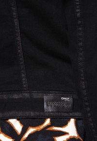 Giacca in denim nera con una texture liscia, caratterizzata da cuciture a contrasto e un'etichetta contrassegnata "ONLY". Sullo sfondo si trova un motivo floreale colorato.