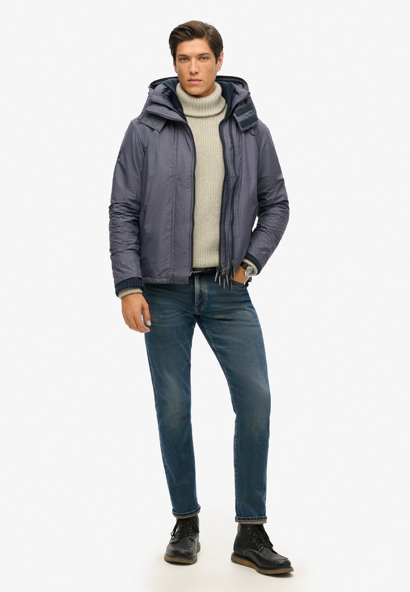 Lekka męska kurtka z kapturem w stonowanej szarości, pod spodem gruby sweter z golfem, w połączeniu z niebieskimi dżinsami i czarnymi butami.