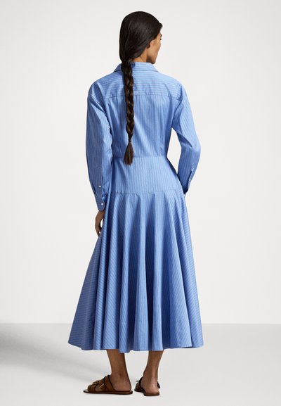 Ženska z dolgo kitko, oblečena v modro obleko z dolgimi rokavi in rjavimi ravnimi sandali, stoji obrnjena stran na enobarvnem ozadju.