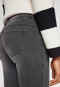 Gray denim jeans with a fitted design, featuring two back pockets and subtle stitching details. Part of a textured, striped sweater is visible.