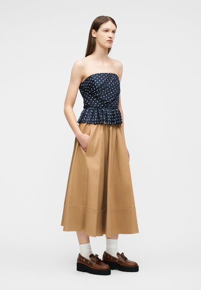 Γυναίκα που φοράει μπλουζάκι χωρίς τιράντες μπλε ναυτικό με πουά, καφέ μεσαίο φούστα με τσέπες, λευκές κάλτσες και καφέ πλατφόρμα μοκασίνια, στέκεται μπροστά σε απλό φόντο.