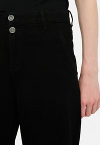 Pantaloni din denim negri cu talie înaltă, având două nasturi metalici în partea de sus, buzunare laterale și o textură netedă.