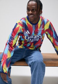 Långärmad, flerfärgad sporttröja med en abstrakt design. Tillverkad av tyg, med blå detaljer och en vit logotyp. Bärs tillsammans med blå jeans.