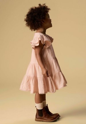 Ragazza giovane con capelli ricci che indossa un vestito rosa chiaro con volant, calzini crema e stivali di pelle marroni, in piedi di profilo su uno sfondo beige.