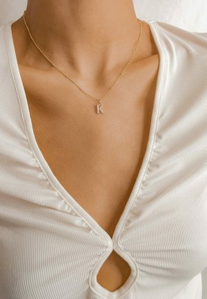 Collier en chaîne en or avec une surface texturée et un pendentif en forme de 'K' incrusté de diamants, porté par-dessus un haut à côtes blanc avec un décolleté en V profond et des coutures décoratives.
