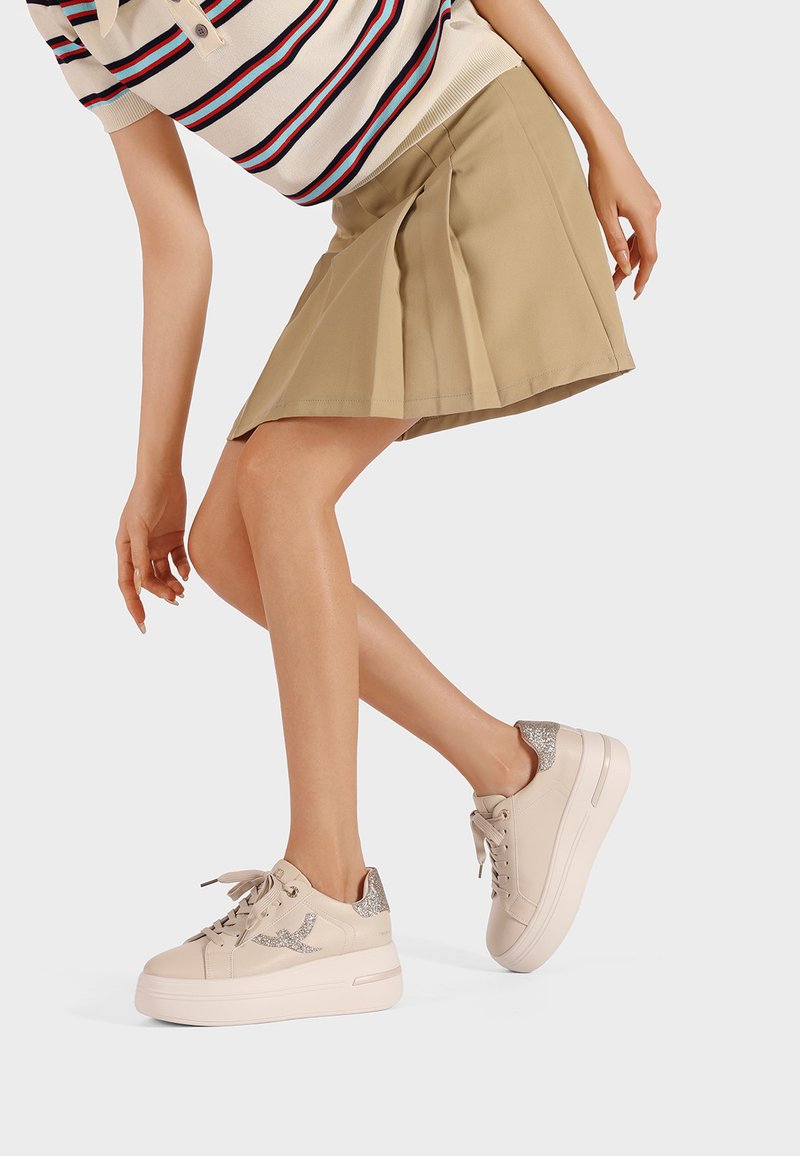 Beżowe trampki na platformie z błyszczącymi srebrnymi akcentami, połączone z beżową plisowaną spódnicą. Projekt charakteryzuje się gładką teksturą i okrągłym przodem.
