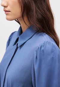 Μπλε μπλούζα με γιακά, μαλακά πτυχώματα στις μανίκες και λεία υφή, αποτυπωμένη από πλάγια γωνία που προβάλλει τον σχεδιασμό της.