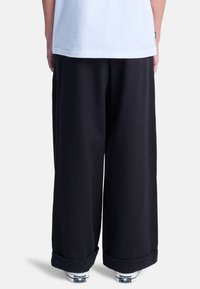 Schwarze weit geschnittene Sweatpants mit weicher Textur, ausgestattet mit umgeschlagenen Säumen. Tragen mit weißen Socken und Leinenschuhen. Rückansicht.