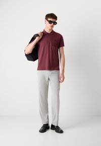 Bordová polokošeľa s vreckom, svetlošedé upravené nohavice, čierne loaferové topánky a slnečné okuliare, doplnené čiernou taškou.