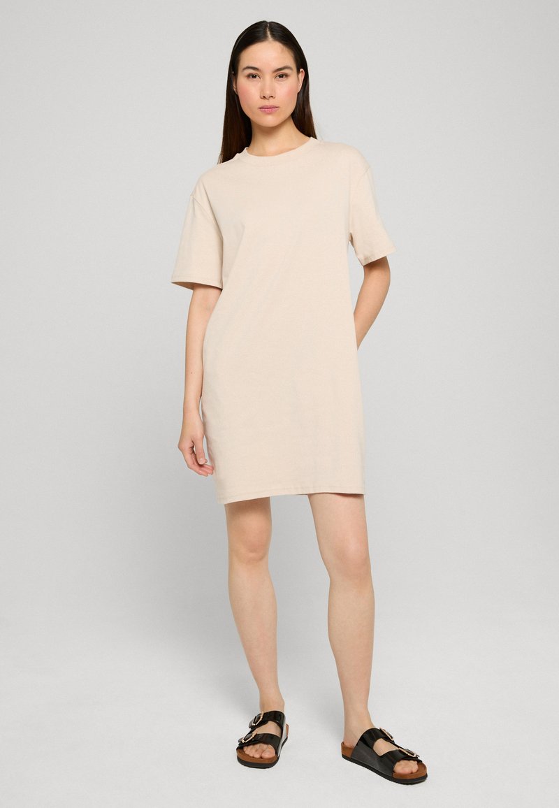 Femme debout portant une robe beige courte à manches courtes et des sandales à boucle noires sur un fond clair uni.