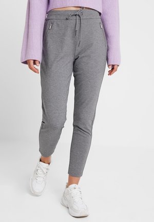 Pantalones de chándal grises con cintura elástica, cordón y dos bolsillos con cremallera. Textura suave y corte ajustado, combinados con zapatillas blancas.