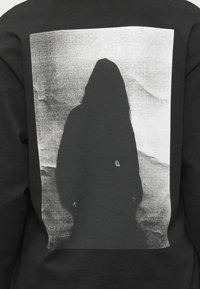 Melns džemperis ar lielu baltu grafiku, kurā attēlots siluets ar gariem matiem uz teksturēta fona. Minimālisma dizains.