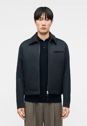 Ung mand iført en sort jakke med lynlås over en mørk skjorte og beige bukser, stående mod en ensfarvet lys baggrund.