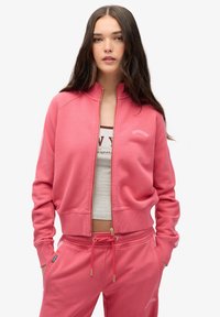 Sudadera rosa con cremallera, cuello y puños acanalados, con la palabra "SUPERDRY" bordada. Combinada con pantalones deportivos rosa a juego.