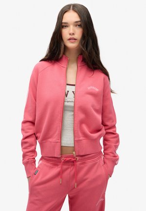 Sudadera rosa con cremallera, cuello y puños acanalados, con la palabra "SUPERDRY" bordada. Combinada con pantalones deportivos rosa a juego.