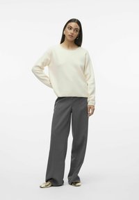 Un sweat-shirt doux de couleur crème avec un col rond, associé à un pantalon gris ample et plissé ainsi qu'à des chaussures plates rondes dorées.