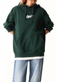 Hoodie vert oversize fabriqué dans un tissu à finition texturée. Doté d'une poche kangourou et d'une broderie de logo blanche sur la poitrine. Associé à un jean clair.