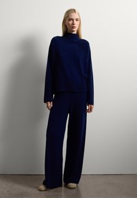 Femme blonde se tenant contre un mur blanc, portant un pull à col roulé bleu marine, un pantalon large assorti et des chaussures beiges.