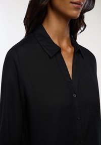 Czarna koszula zapinana na guziki z kołnierzykiem, gładką teksturą i lekko dopasowanym krojem. Skupienie na detalu listwy guzikowej i guzików.