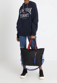 Borsa tote nera con manici rossi e blu, ferrature argentate e una targhetta. Materiale morbido, forma rettangolare e design funzionale.