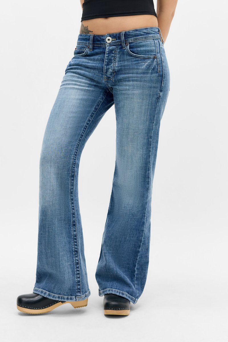 Άτομο που φοράει ξεθωριασμένο μπλε τζιν bootcut και μαύρα ξύλινα παπούτσια με ξύλινη σόλα, στέκεται μπροστά σε απλό φόντο.