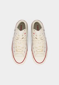 Εκρού καμβαδένια αθλητικά παπούτσια ψηλής κορυφής με λευκά κορδόνια, κόκκινη επένδυση στις σόλες, μεταλλικές οπές και διακριτικά μεταλλικά τρουκ σε σχήμα αστεριού στα πλάγια.