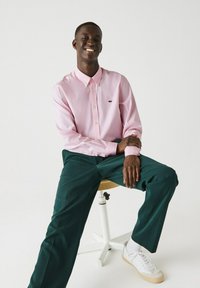 Camicia rosa a maniche lunghe con colletto aperto, abbinata a pantaloni verde scuro e sneakers bianche. Seduto su uno sgabello contro uno sfondo bianco.