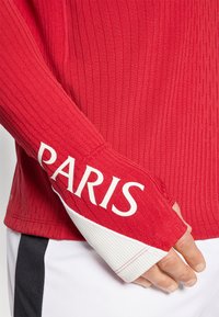 Piros, hosszú ujjú felső textúrált anyagból, fehér díszítéssel az ujjon, amelyen vastag betűkkel „PARIS” felirat található.