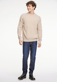 Beiger Strickpullover mit langen Ärmeln, kombiniert mit dunkelblauen Jeans und schwarzen Stiefeln. Der Pullover hat einen runden Halsauschnitt und gerippte Bündchen.