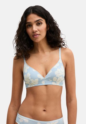 Femme aux cheveux bouclés et foncés portant un soutien-gorge triangle fleuri bleu clair et une culotte taille basse assortie, sur un fond uni.