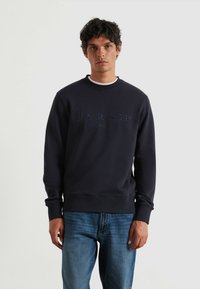 Marine crewneck sweatshirt met geborduurd merkl logo, geribbelde manchetten en onderkant, gemaakt van zachte stof, gedragen over blauwe jeans.