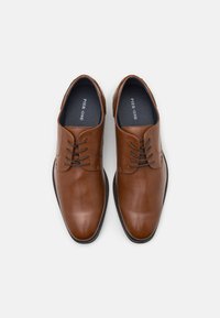 Zapatos de vestir de cuero marrón con un acabado suave, punta redondeada, cierre con cordones y suela negra. El interior presenta la etiqueta de la marca.