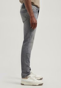 Karcsú szabású szürke farmernadrág, megfakult kivitelben, öt zsebbel és logós toldással a hátoldalon. Fehér sportcipővel párosítva.