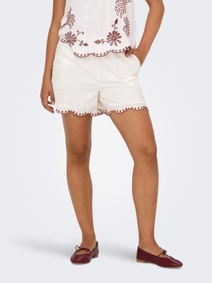 Kvinde iført hvide shorts med bølget kant, brune broderede prikker og brune balletsko, står med den ene hånd i lommen.