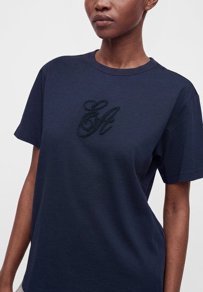 Γυναίκα που φοράει μπλε σκουρόχρωμο t-shirt με κοντά μανίκια και υφασμένα μαύρα γράμματα "St" στο στήθος.