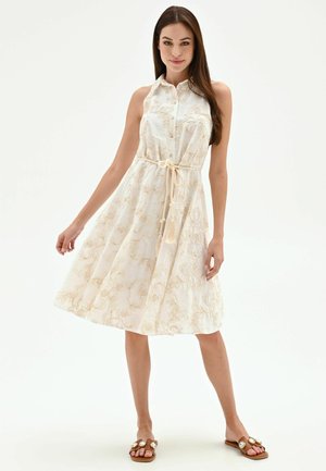 Femme portant une robe blanche sans manches avec broderies florales, une ceinture nouée à la taille et des sandales marron avec de grosses pierres décoratives.
