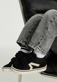 Svarta sneakers med två kardborreband, vita detaljer och en strömlinjeformad design. Bärs tillsammans med grå denimbyxor och randiga strumpor.