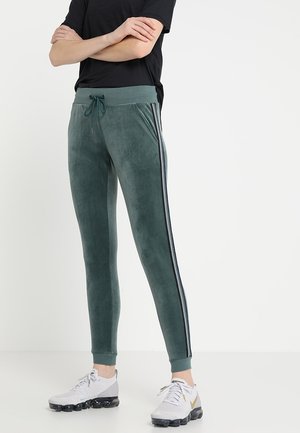 Person iført grønne velour joggebukser med sort-hvide sidelister, sort t-shirt med korte ærmer og hvide sportssko med sorte såler.