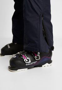 Sorte skistøvler med metalspænder og lilla accenter. Navy skibukser med lynlås ved ankelen, der viser støvlens design detaljer.