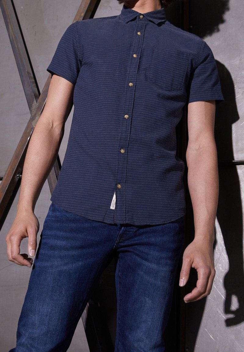 Camicia blu scuro a maniche corte con un motivo testurizzato, chiusura con bottoni, taschino e un'etichetta all'orlo, abbinata a jeans di denim blu.