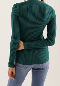 Femme portant un pull côtelé ajusté vert foncé et un jean bleu clair, vue de dos avec une main derrière le dos.