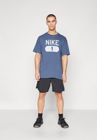Blå bomulds T-shirt med hvid "NIKE 1" print, kombineret med sorte sportshorts og sorte sneakers, stående mod en ensfarvet baggrund.