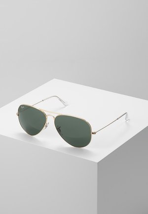 Lunettes de soleil aviateur avec une monture en métal doré et des verres verts, dotées d'un pont double et d'accents argentés sur une base blanche.