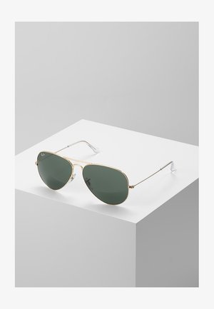 Arany fémkeretes pilóta napszemüveg zöld lencsékkel, dupla híd jellemzi, ezüst szárakkal egy fehér talapzaton.