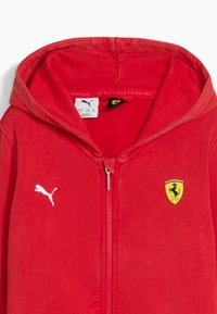 Rotes Kapuzensweatshirt mit einem Frontreißverschluss, versehen mit einem weißen Puma-Logo und einem gelben Ferrari-Schild-Logo. Weicher Stoff mit glatter Textur.