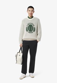 Maglione bianco lavorato a maglia con logo verde "LACOSTE PARIS", pantaloni neri, sneakers bianche e una borsa crema con accenti neri.